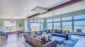 A spacious living room with a large sectional sofa, a coffee table, and a dining area with a wooden table and chairs. Large windows provide a view of the beach.