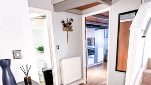 Käytävän lattia on parkettia, siellä on musta maljakko ja patteri. Portaat, joiden päällä on beigenvärinen matto, johtavat toiseen huoneeseen.
