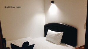 Habitación semi-privada con una cama individual, una almohada, una toalla doblada y una mesita de noche.