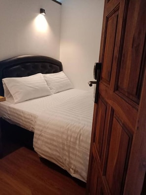 A bedroom with a bed, a nightstand, a wooden door, and a wall-mounted light. - Seahorse Lipe Hostel (Koh Lipe)