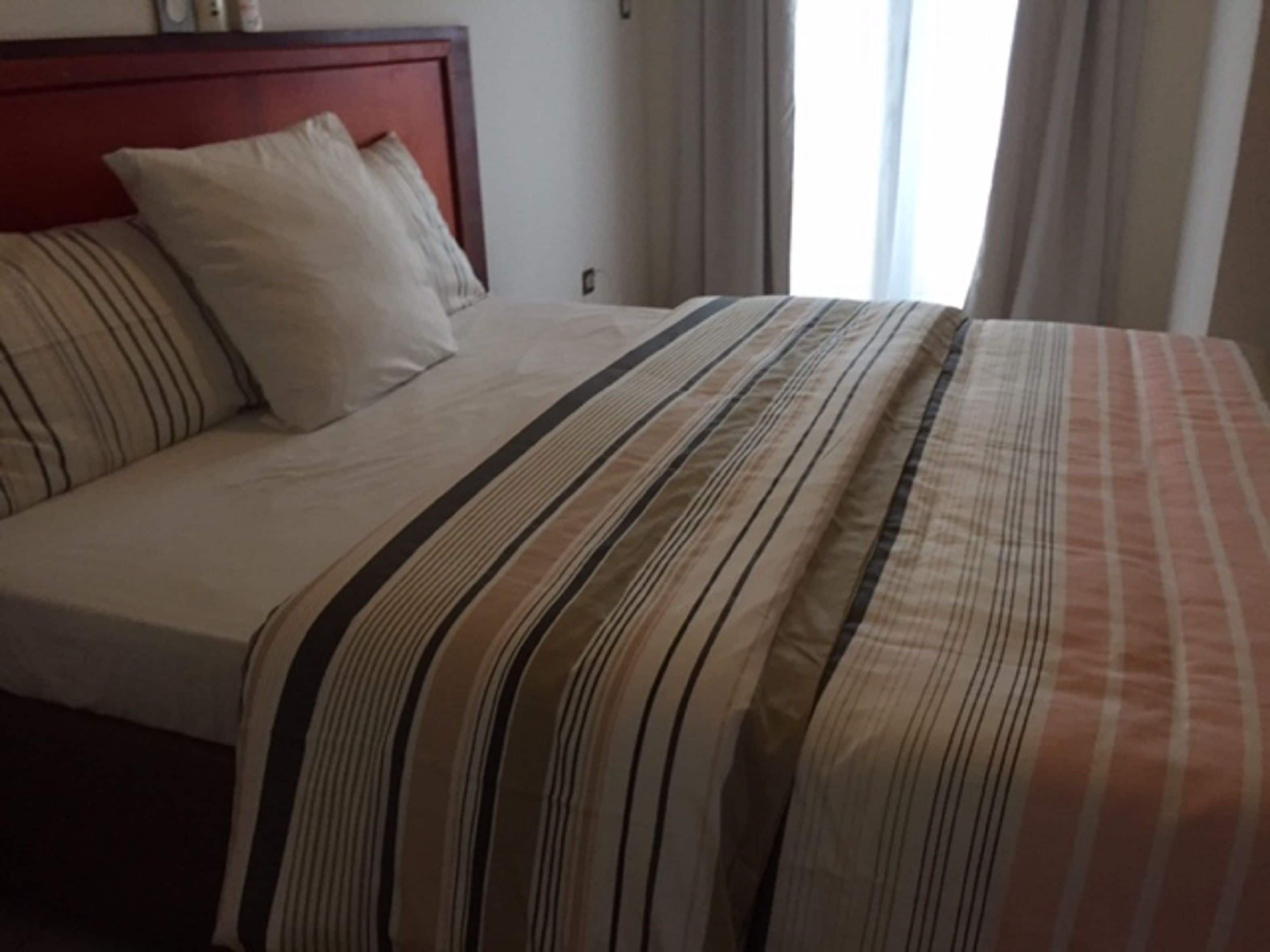 A neatly made bed with striped bedding, a wooden headboard, and a plain wall with a small clock.