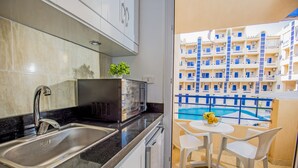 Et køkken med vask, mikroovn og en potteplante. Der er en balkon med pool og et bord med stole.