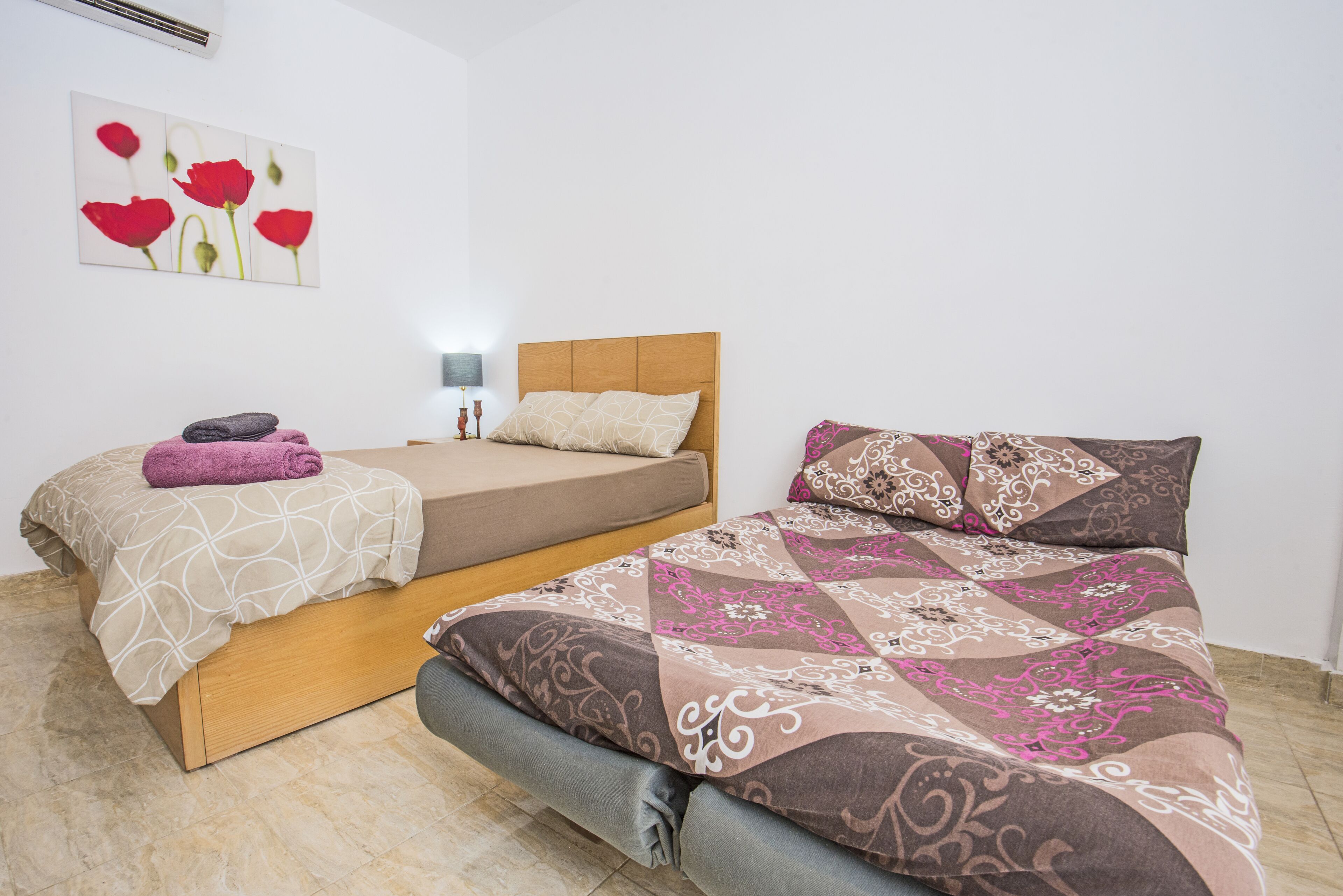 Une chambre avec deux lits, un climatiseur fixé au mur, une lampe de chevet et un tableau représentant des fleurs rouges.