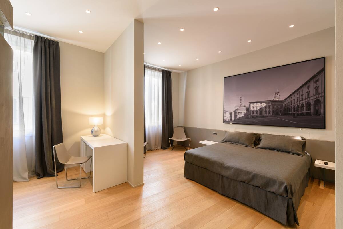 Ett modernt hotellrum med en stor säng, ett skrivbord med en stol, ett litet bord och en stor, inramad tavla på väggen.