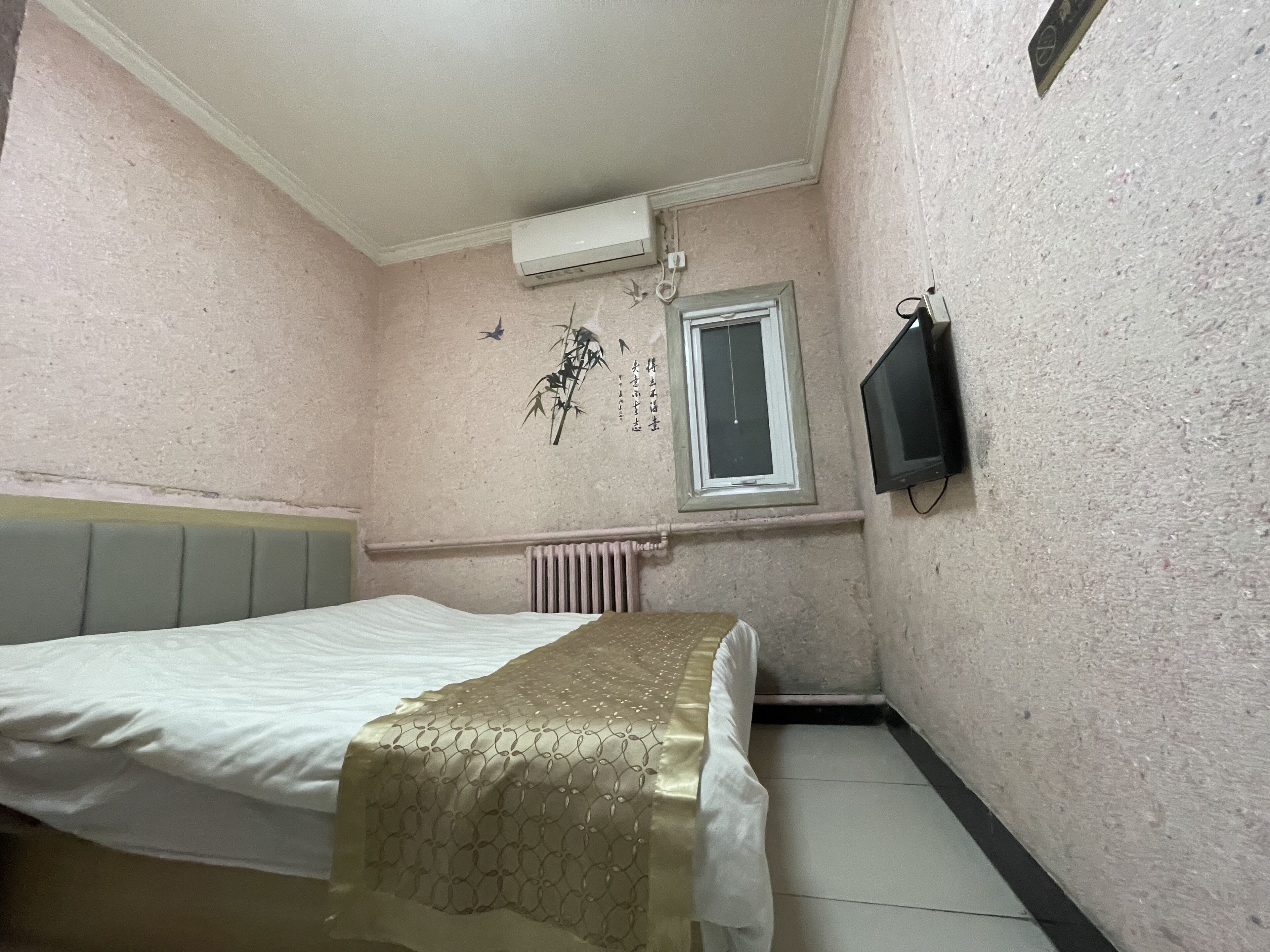 Una habitación pequeña y luminosa con cama, radiador, ventana y televisor montado en la pared.