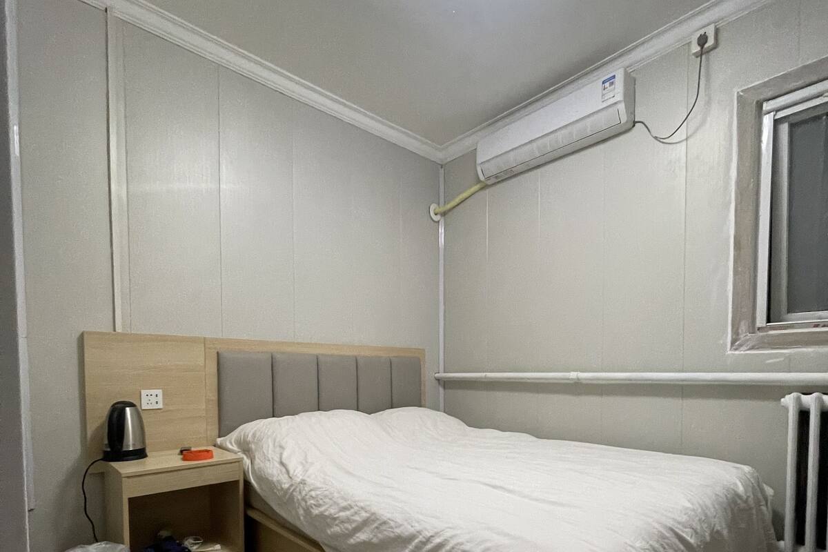 Une petite chambre avec un lit simple, une tête de lit en bois, une table de chevet avec une bouilloire et un radiateur.