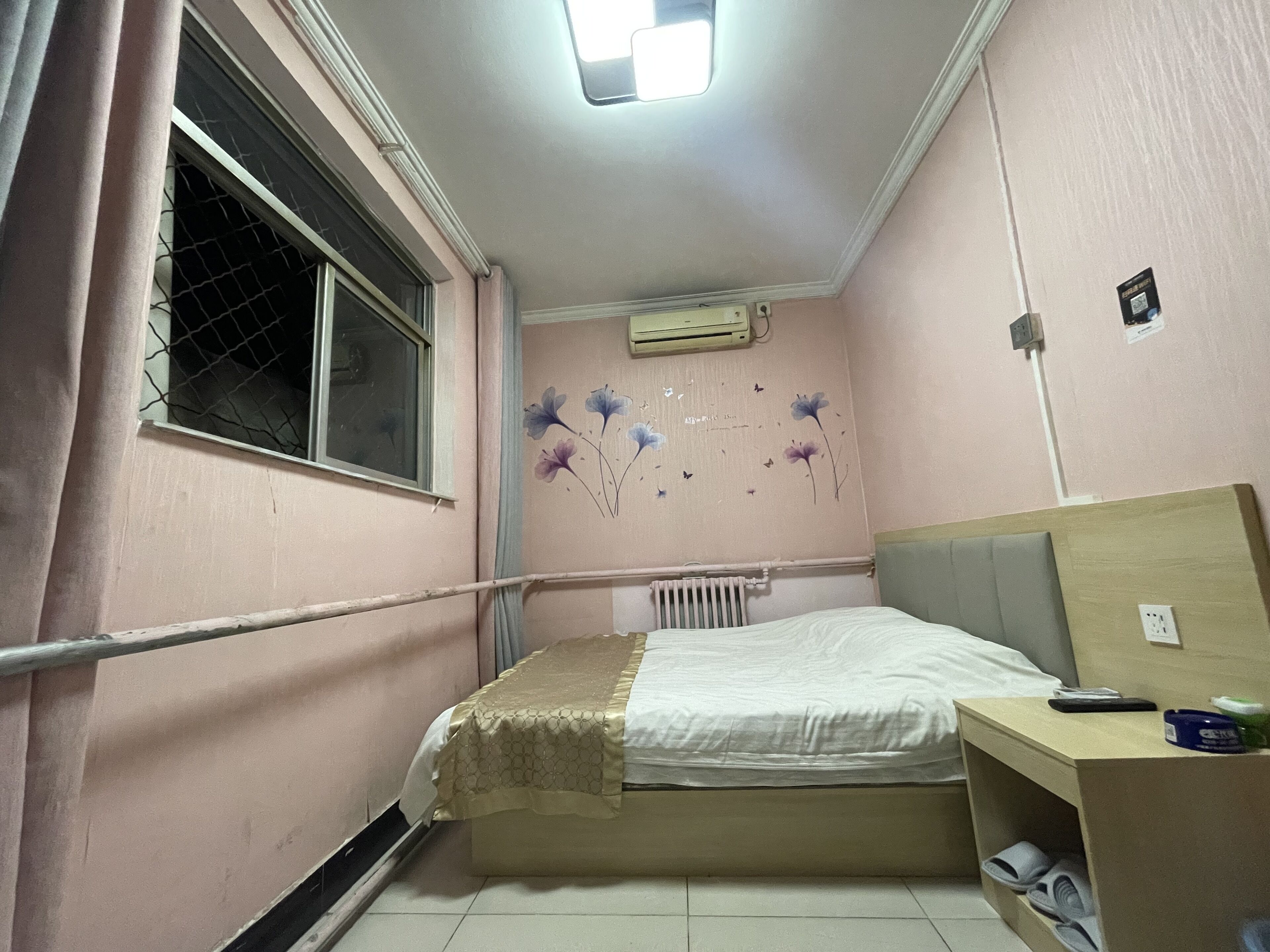 A small, pink-walled room with a single bed, a radiator, a window with a metal screen, and a small desk with a chair.