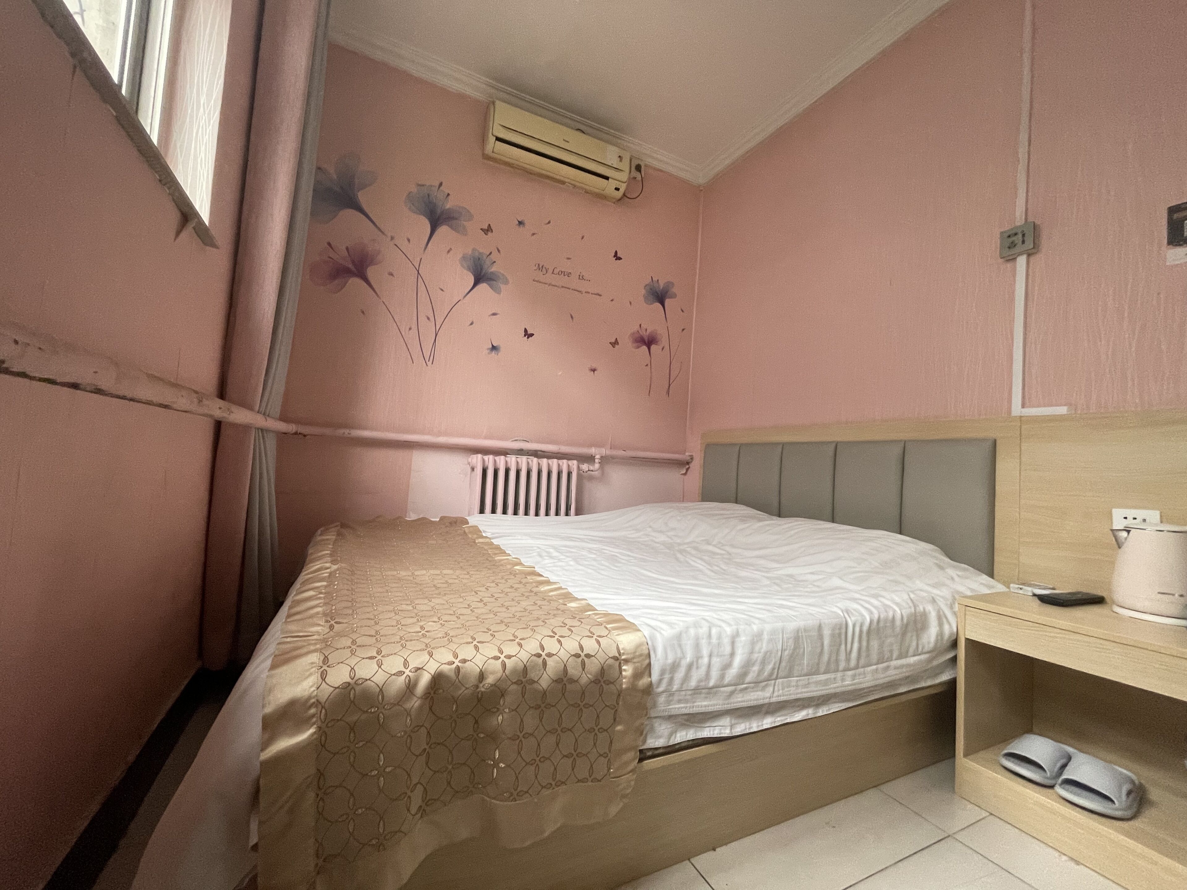 A bedroom with a bed, a radiator, a shelf with slippers, and a wall with floral and butterfly decorations.