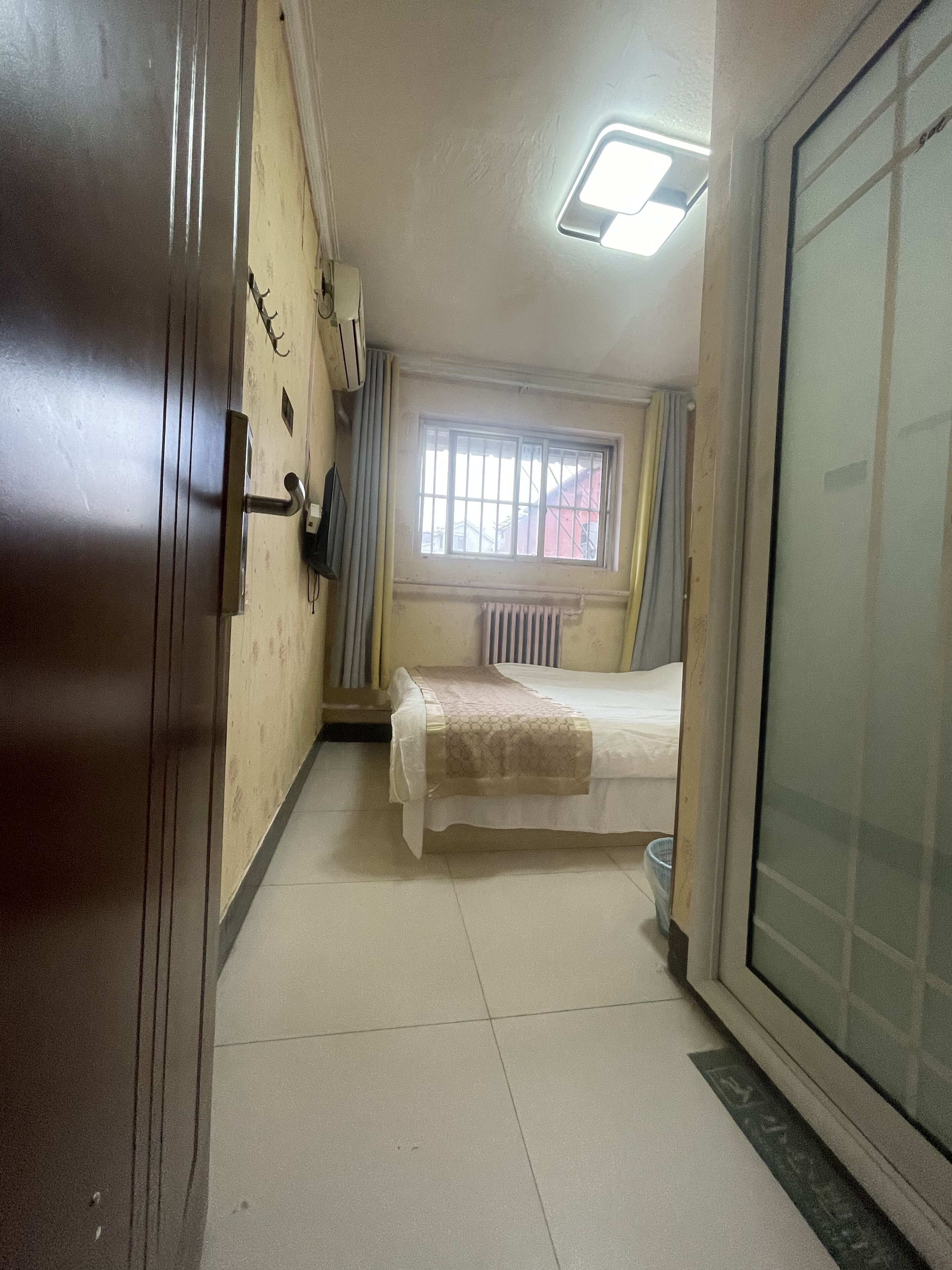 Una habitación pequeña con cama, un radiador, una ventana con rejas y una puerta.