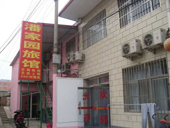A building with a pink exterior and a signboard with yellow text. There are air conditioning units on the wall and a staircase leading to the entrance. - Beijing Huairou Pan Jia Yuan Hotel (Huairou)