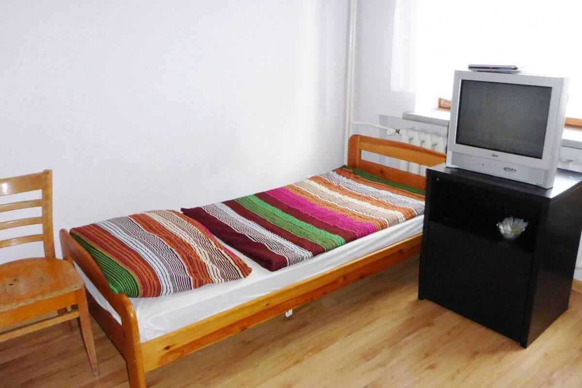 Un lit simple recouvert d’une couverture rayée aux couleurs vives, une chaise en bois, un téléviseur posé sur un meuble et un mur blanc.