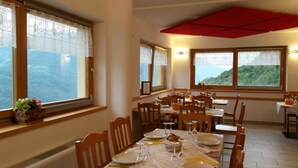 Un comedor con varias mesas y sillas, ventanas amplias con cortinas de encaje y vista al mar.