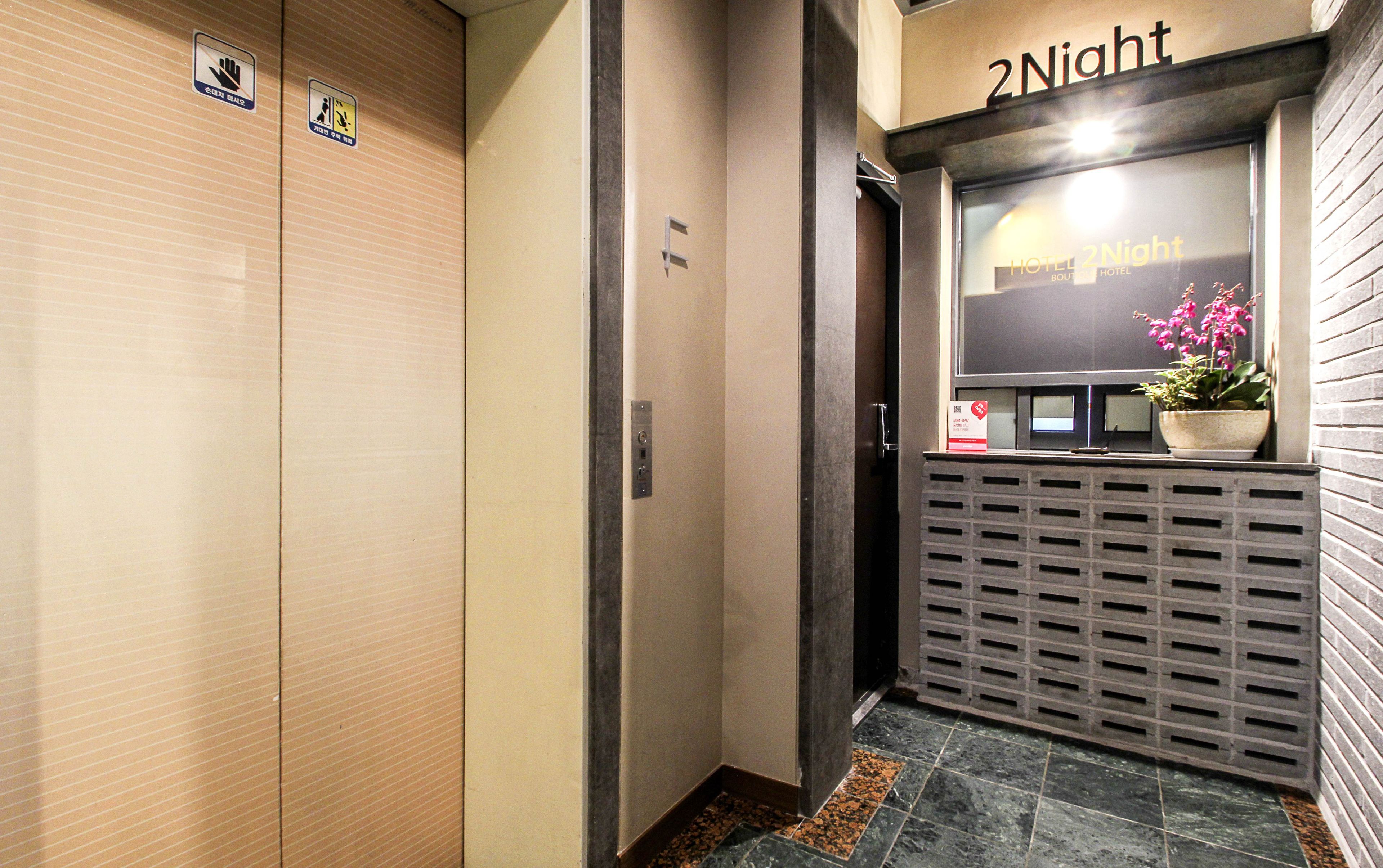 A hotel reception area with a '2Night' sign, a reception desk, a potted plant, and a security sign.