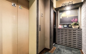Zona de recepción de hotel con un cartel de '2Night', un mostrador de recepción, una planta en maceta y un letrero de seguridad.