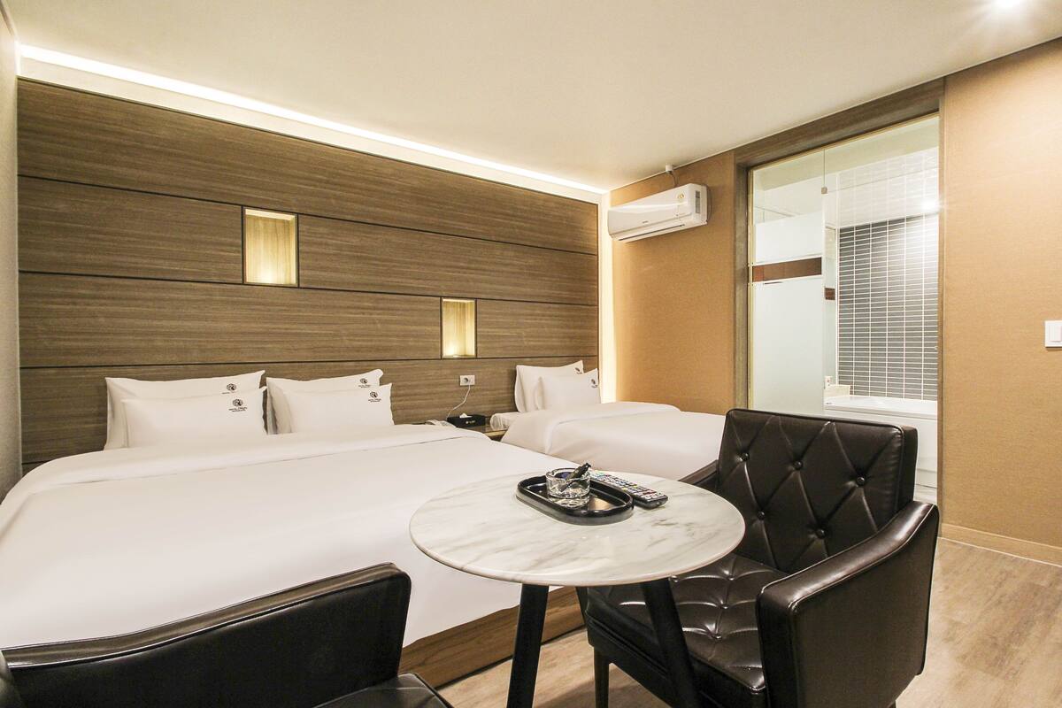 A hotel room with a large bed, a small round table, a leather armchair, and an air conditioning unit on the wall.