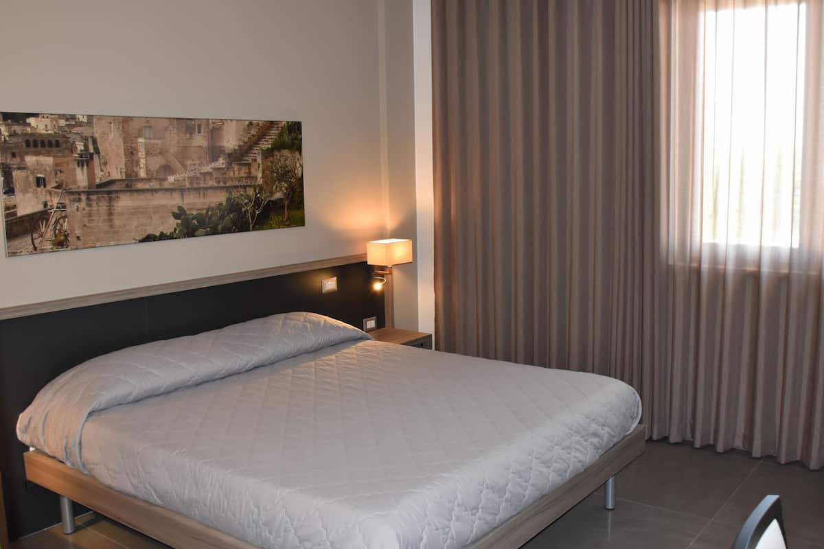 A hotel room with a bed, a nightstand with a lamp, a painting on the wall, and a window with curtains.