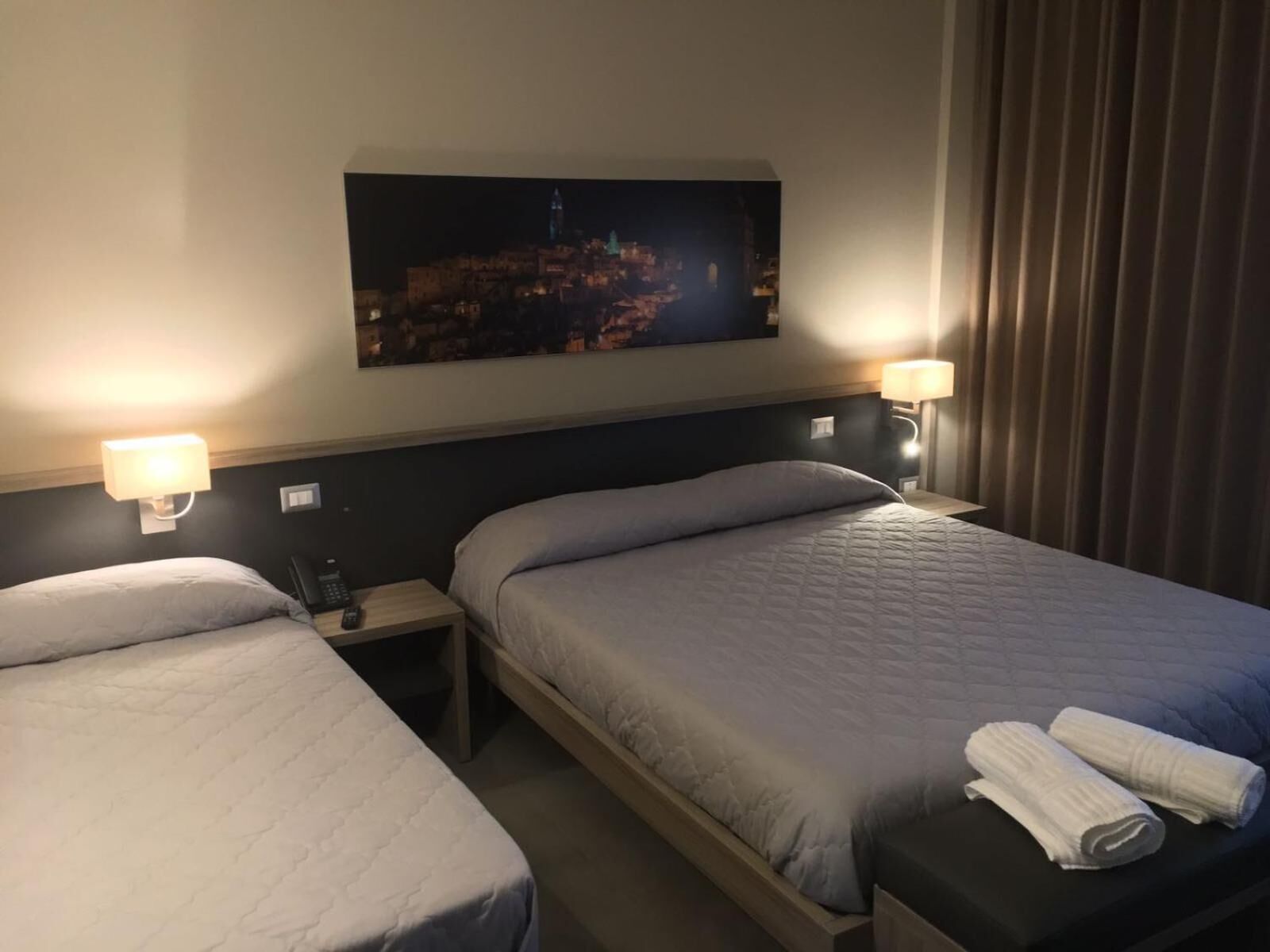 A hotel room with two beds, a nightstand, a lamp, and a painting of a cityscape.