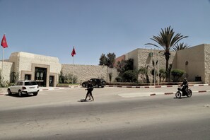 A street with a white car, a motorcycle, and a pedestrian. There are flags on poles, a building with a stone facade, and palm trees. - Hotel Parador (Laayoune)