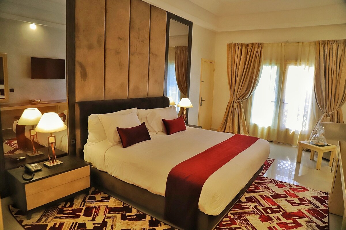 A hotel room with a large bed, two bedside lamps, a wooden headboard, a mirror, a television, and a patterned rug.