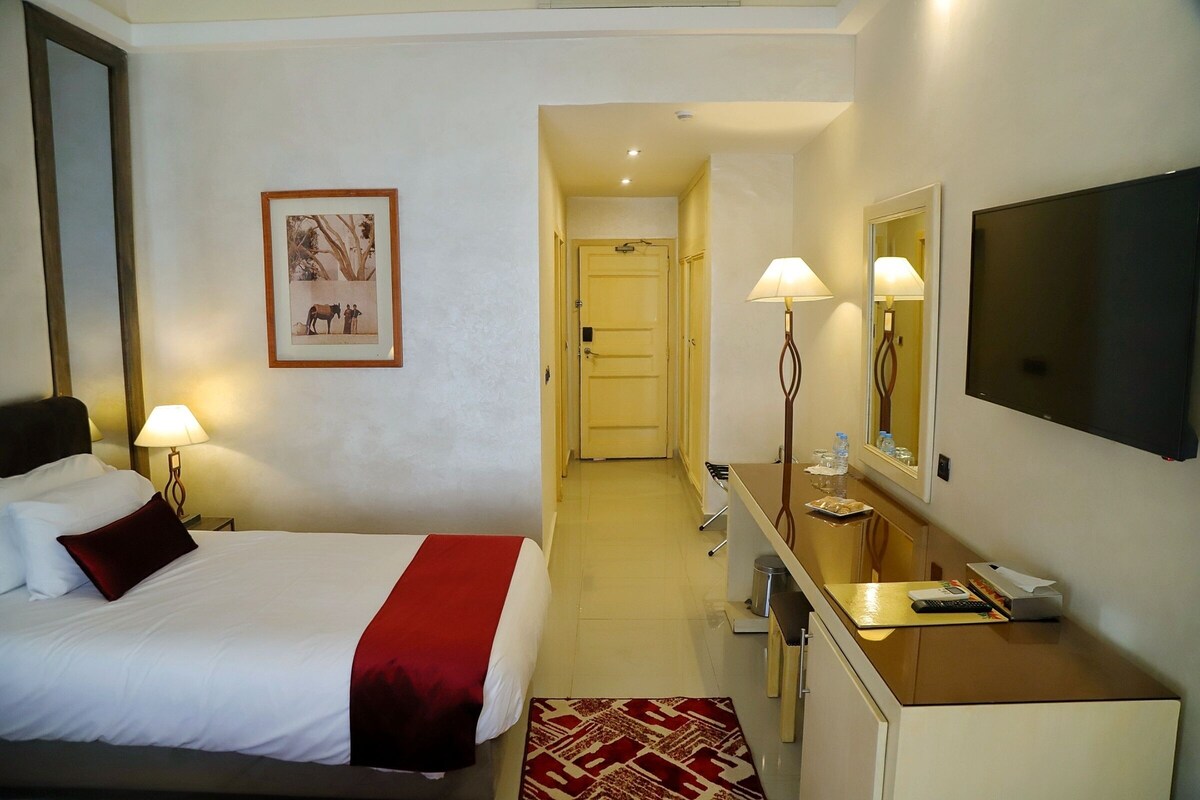A hotel room with a bed, bedside lamps, a TV, a desk with a lamp, a mirror, and a framed picture on the wall.