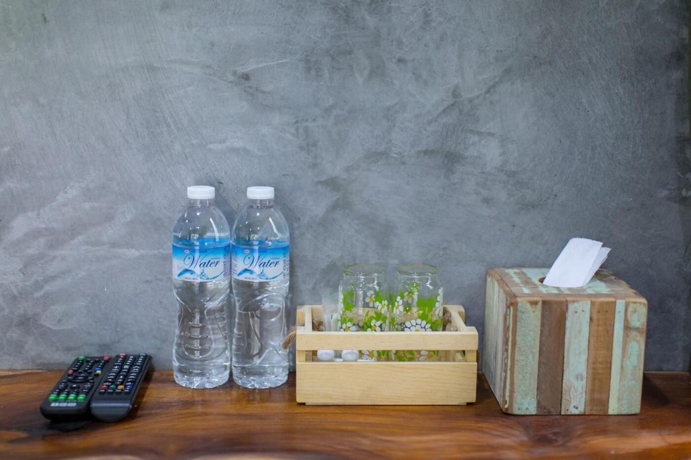 Une télécommande, deux bouteilles d’eau, un bocal en verre avec des fleurs et une boîte de mouchoirs, le tout posé sur une surface en bois devant un mur gris.