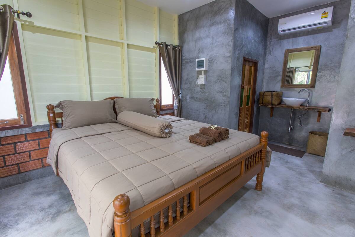 A bedroom with a wooden bed, beige bedding, a small bedside table, a wall-mounted air conditioner, a bathroom with a sink and mirror, and a window with curtains.
