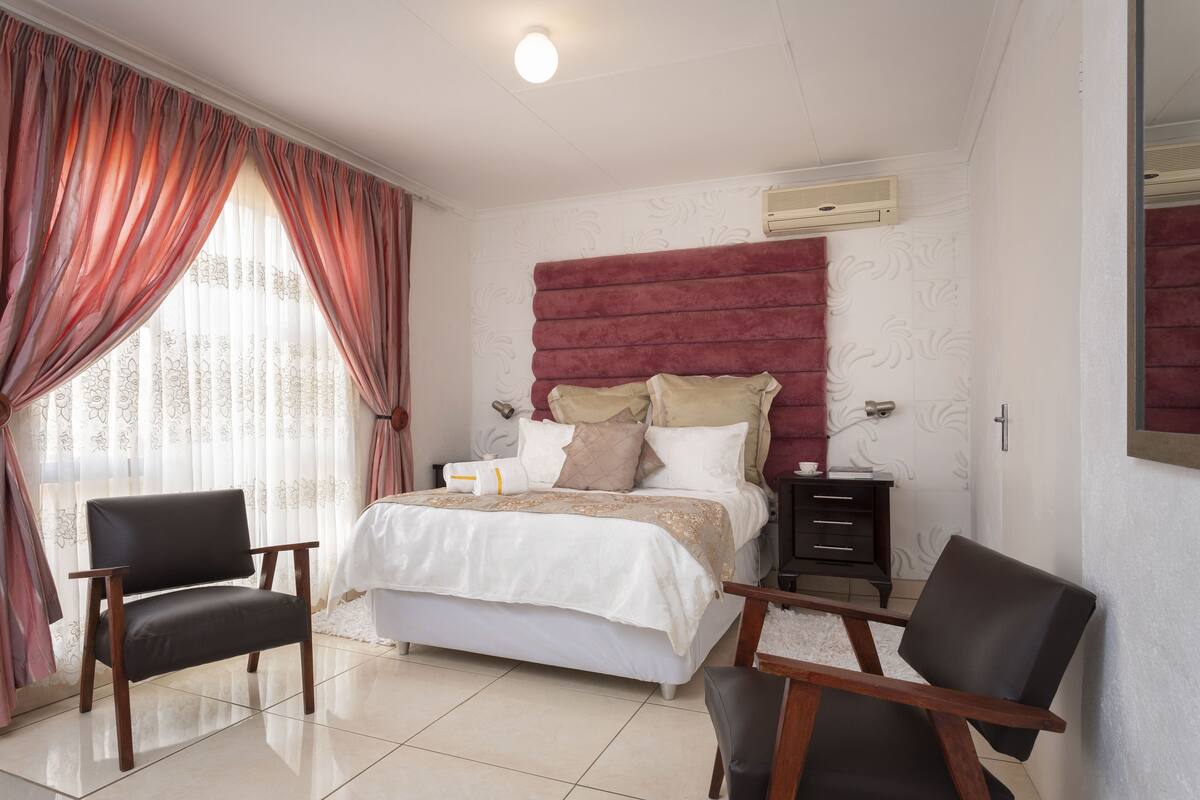 A bedroom with a bed, two chairs, a nightstand, and a window with curtains.