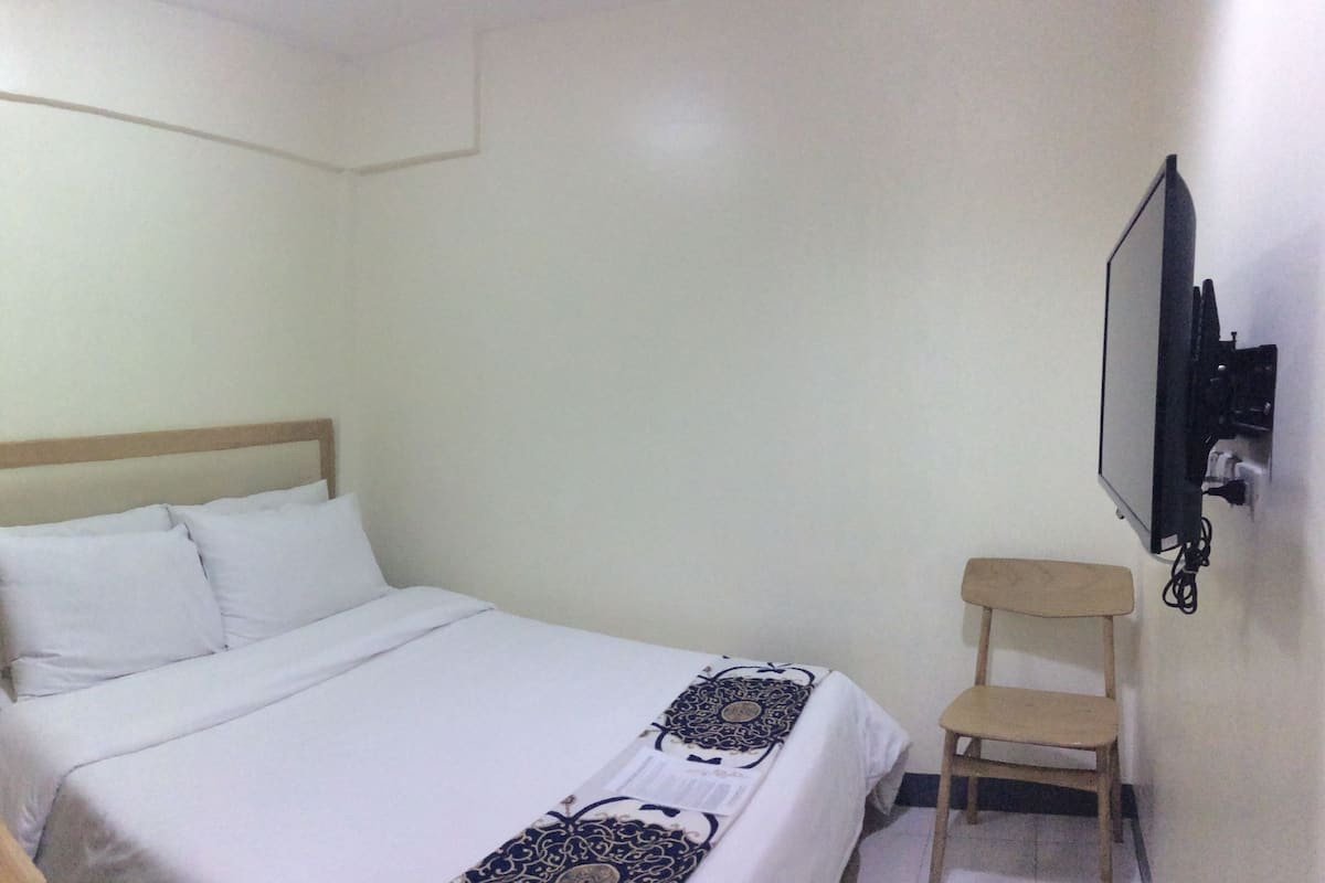 A small, clean room with a single bed, a wooden chair, and a TV mounted on the wall.