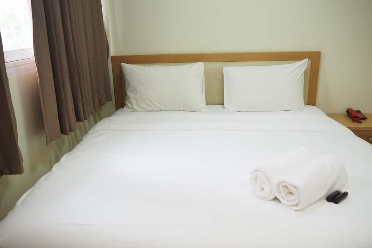 A neatly made bed with white linens and two pillows, a wooden headboard, and a rolled towel on the side.