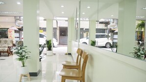 Um hall de entrada limpo e moderno com balcão de recepção, cadeiras de madeira, plantas em vasos e uma parede de vidro com vista para a rua.