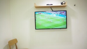Quarto de hotel com uma cama, uma cadeira de madeira, uma televisão de tela plana na parede exibindo uma partida de futebol e uma prateleira acima da TV.