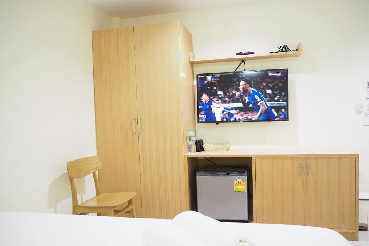 A hotel room with a bed, a wooden wardrobe, a small refrigerator, a television mounted on the wall, and a wooden chair.