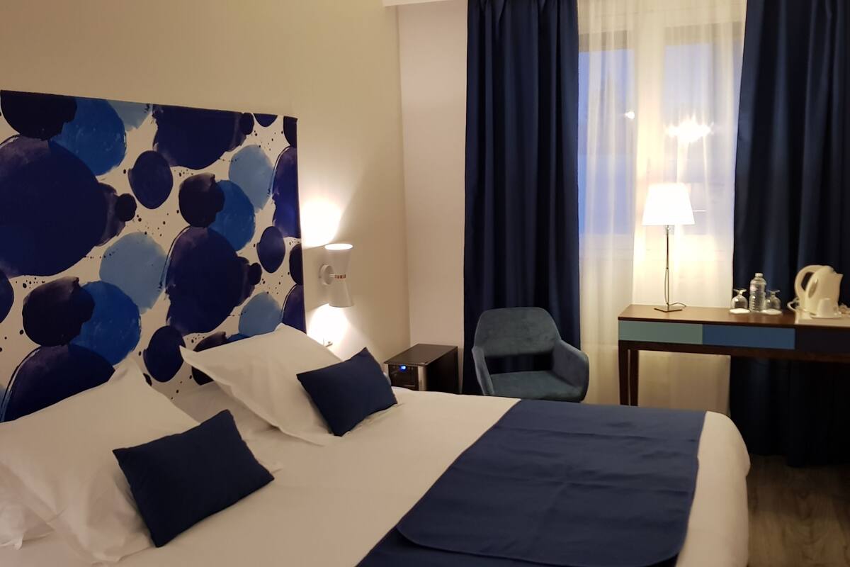 A hotel room with a large bed, a desk with a lamp, a chair, and a TV on the wall.