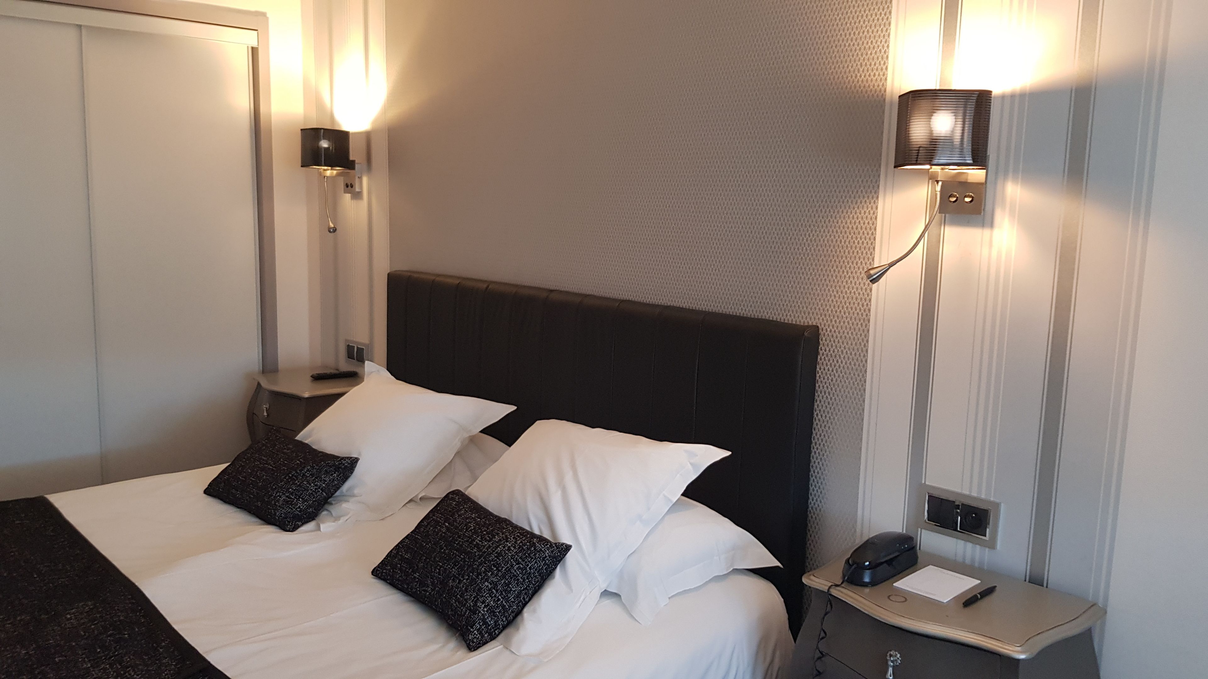 Ein ordentlich gemachtes Bett mit weißen und dunklen Kissen, ein Nachttisch mit Lampe und eine Wandleuchte.