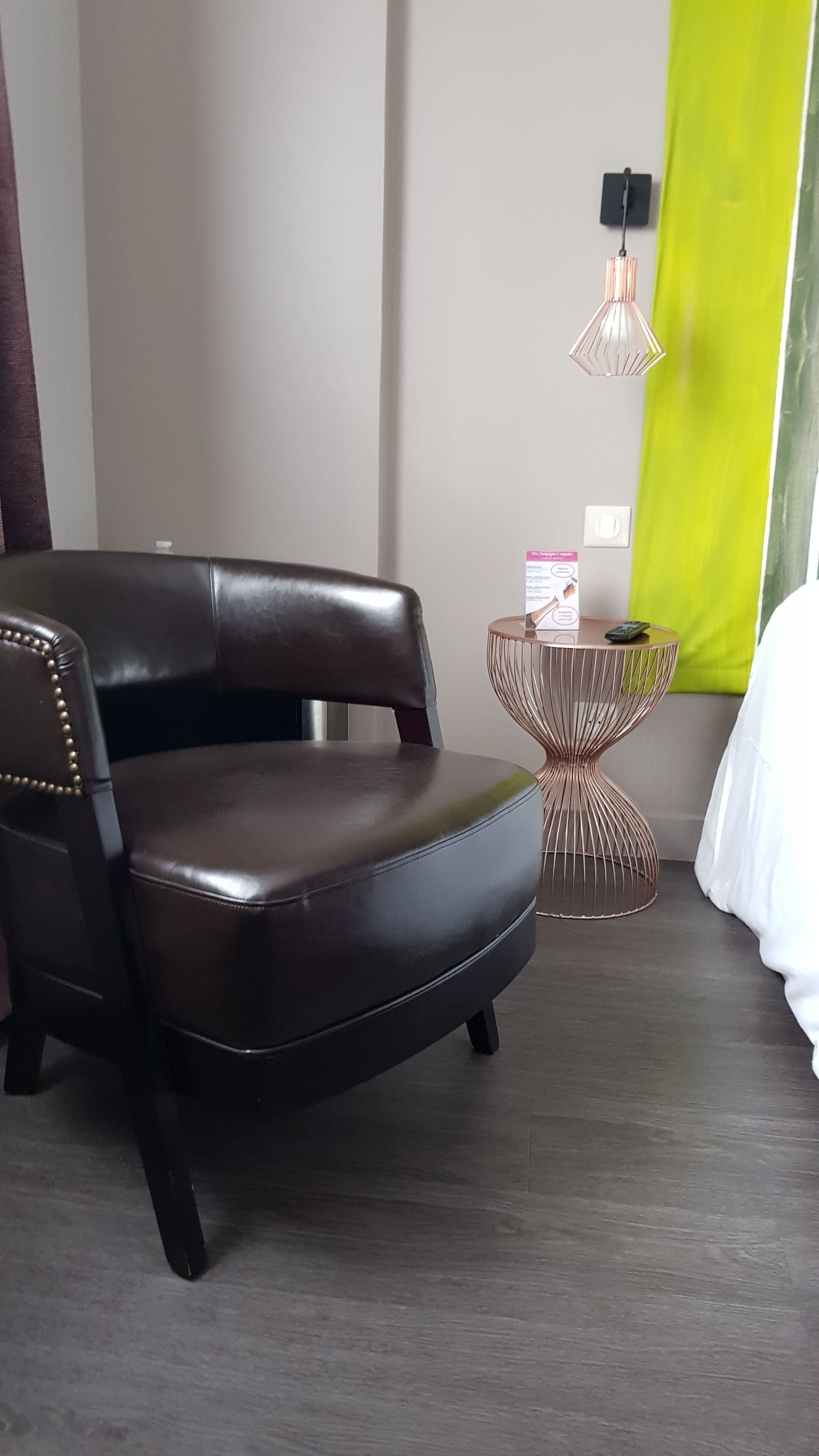 A modern hotel room with a dark brown leather chair, a small round table with a copper base, a green accent wall, and a white bed.