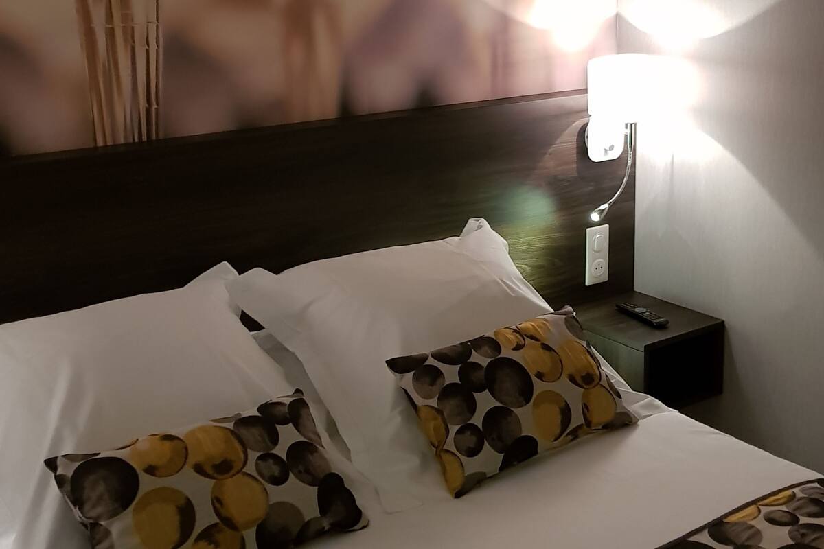 A hotel room with a bed, pillows, a nightstand, a lamp, and a wall with a large abstract painting.