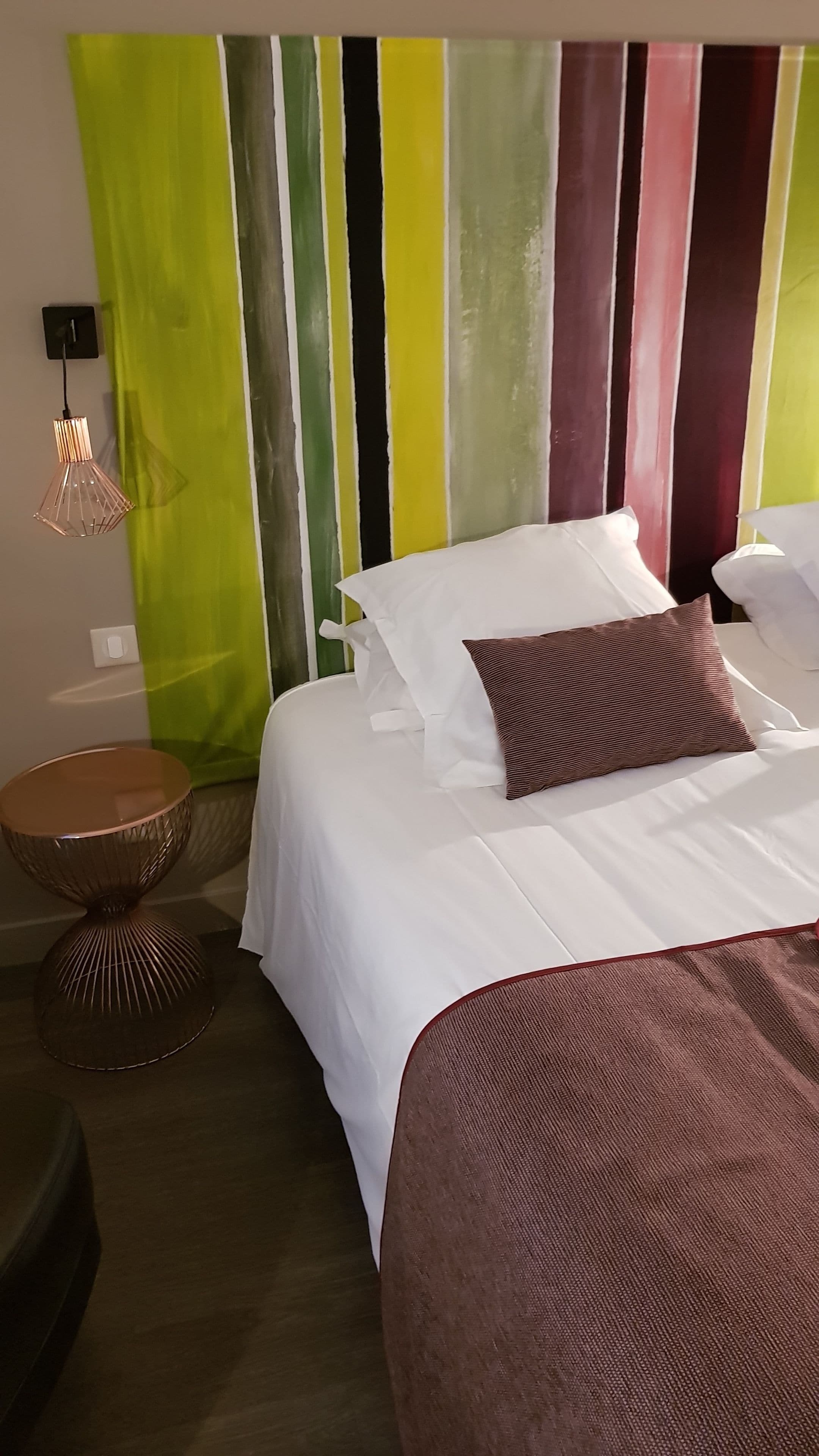 A neatly made bed with white linens and a brown accent pillow, a round wooden side table, and a modern pendant light.
