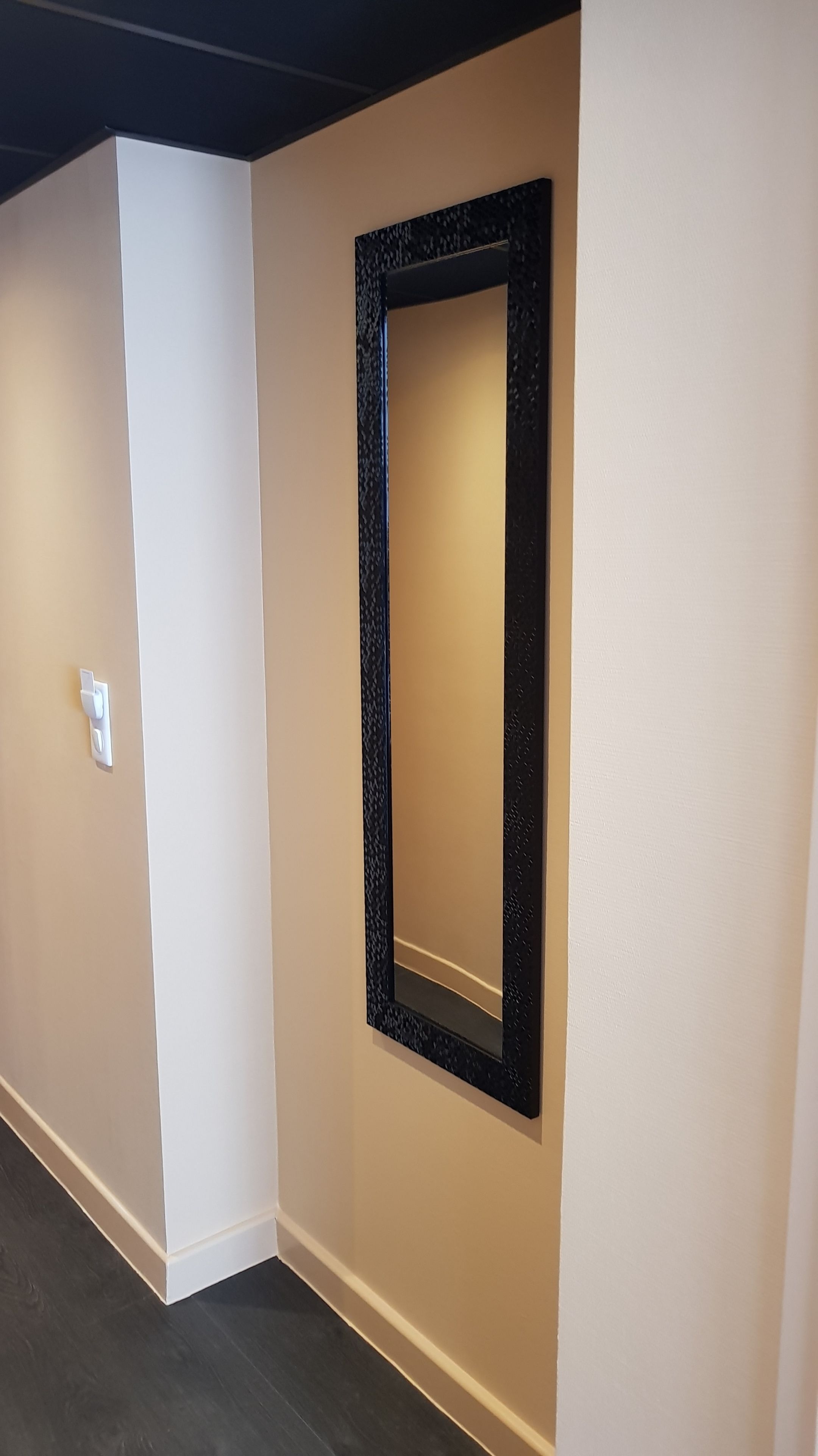 A hallway with a black-framed mirror on the wall, a white switch, and a dark floor.