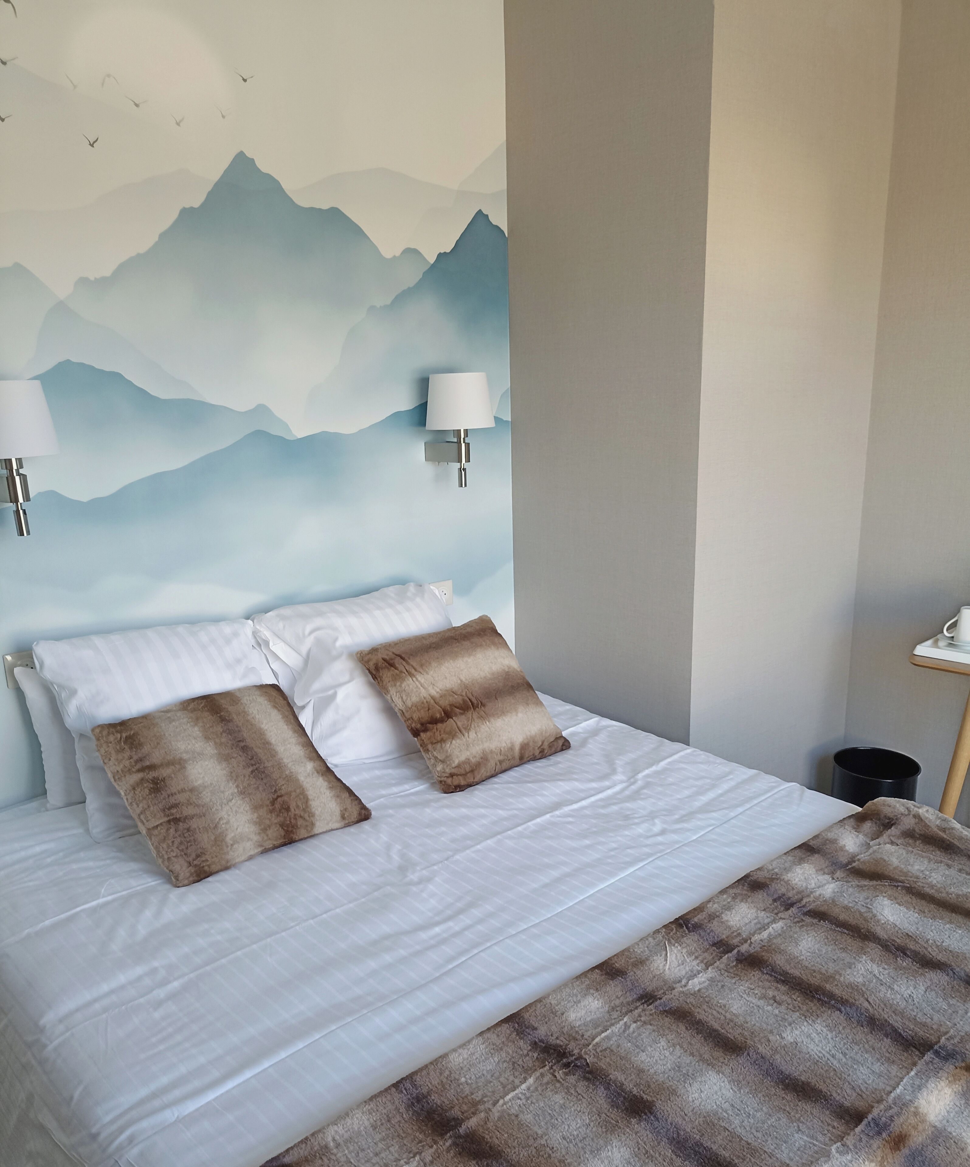 Ein ordentlich bezogenes Bett mit weißen und braunen Kissen, einer Karodecke und einer Wandmalerei von Bergen.