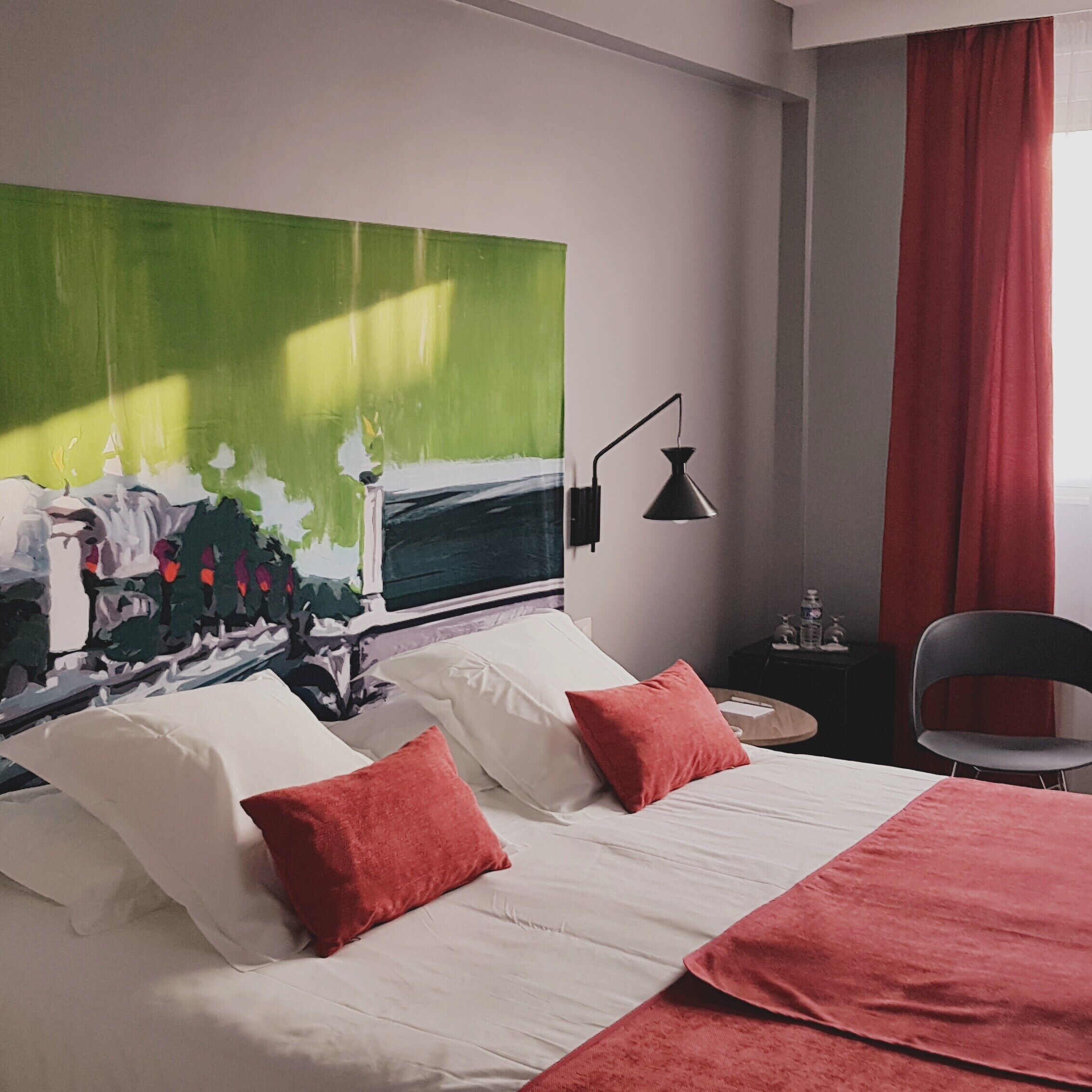 A hotel room with a bed, two pillows, a nightstand, a lamp, a chair, and a large abstract painting on the wall.