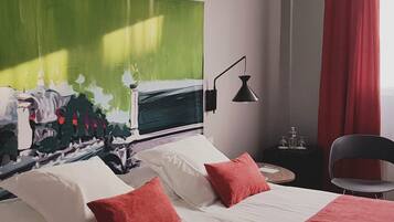A hotel room with a bed, two pillows, a nightstand, a lamp, a chair, and a large abstract painting on the wall.