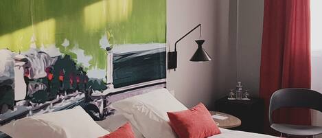 A hotel room with a bed, two pillows, a nightstand, a lamp, a chair, and a large abstract painting on the wall.