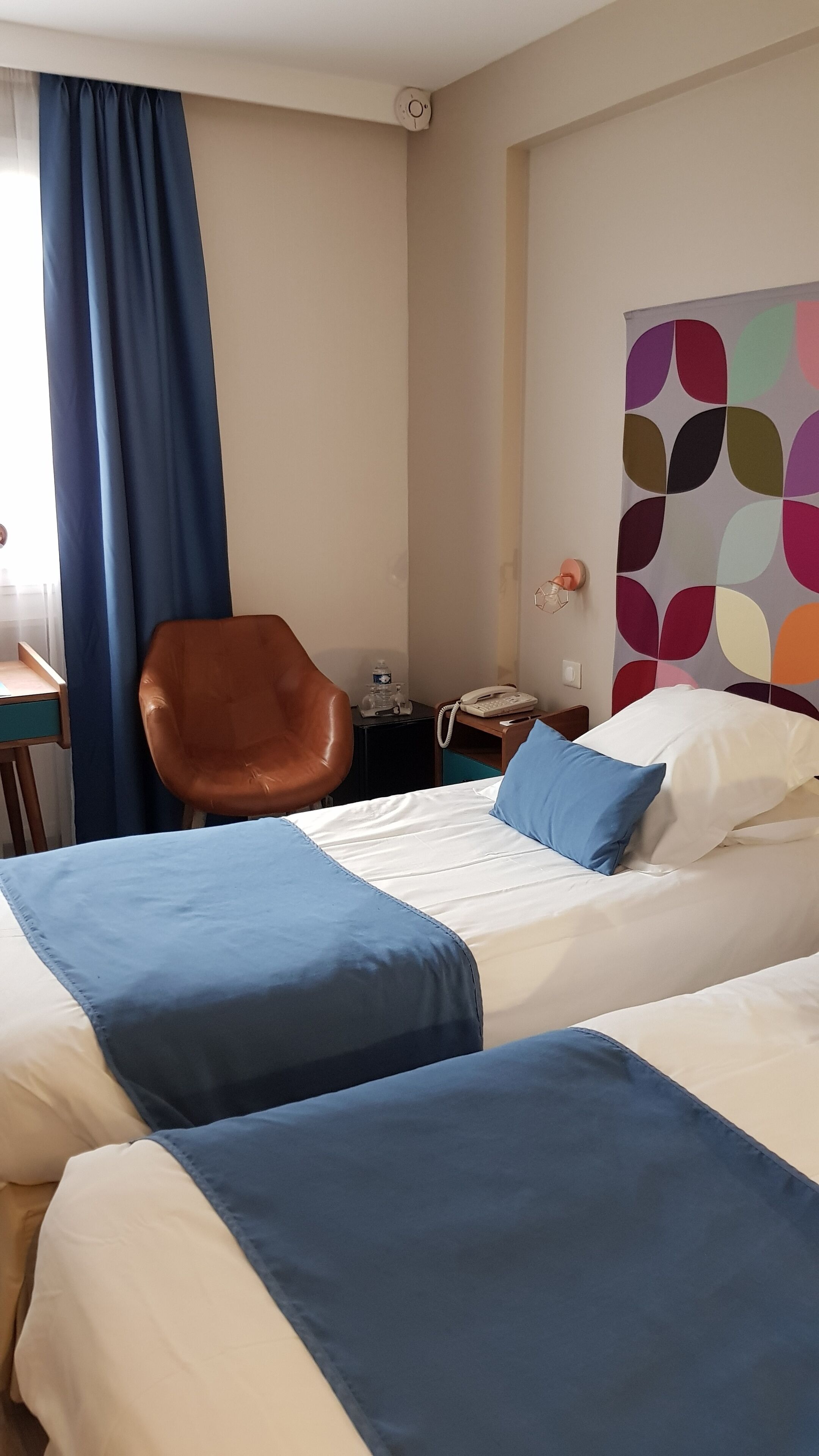 A hotel room with two beds, a desk, a chair, a phone, and a colorful wall decoration.