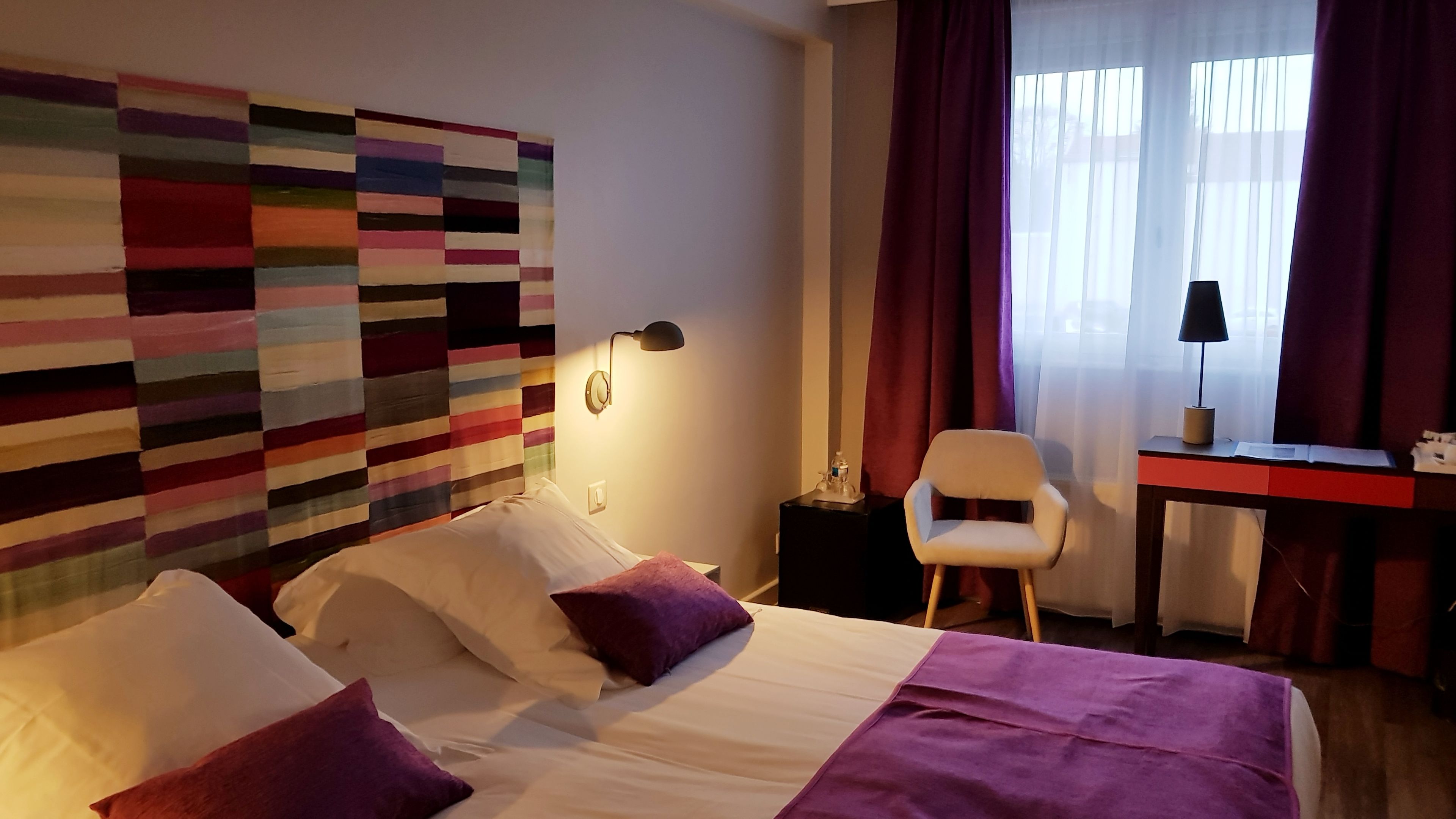 A hotel room with a bed, a desk, a chair, and a colorful wall art piece.