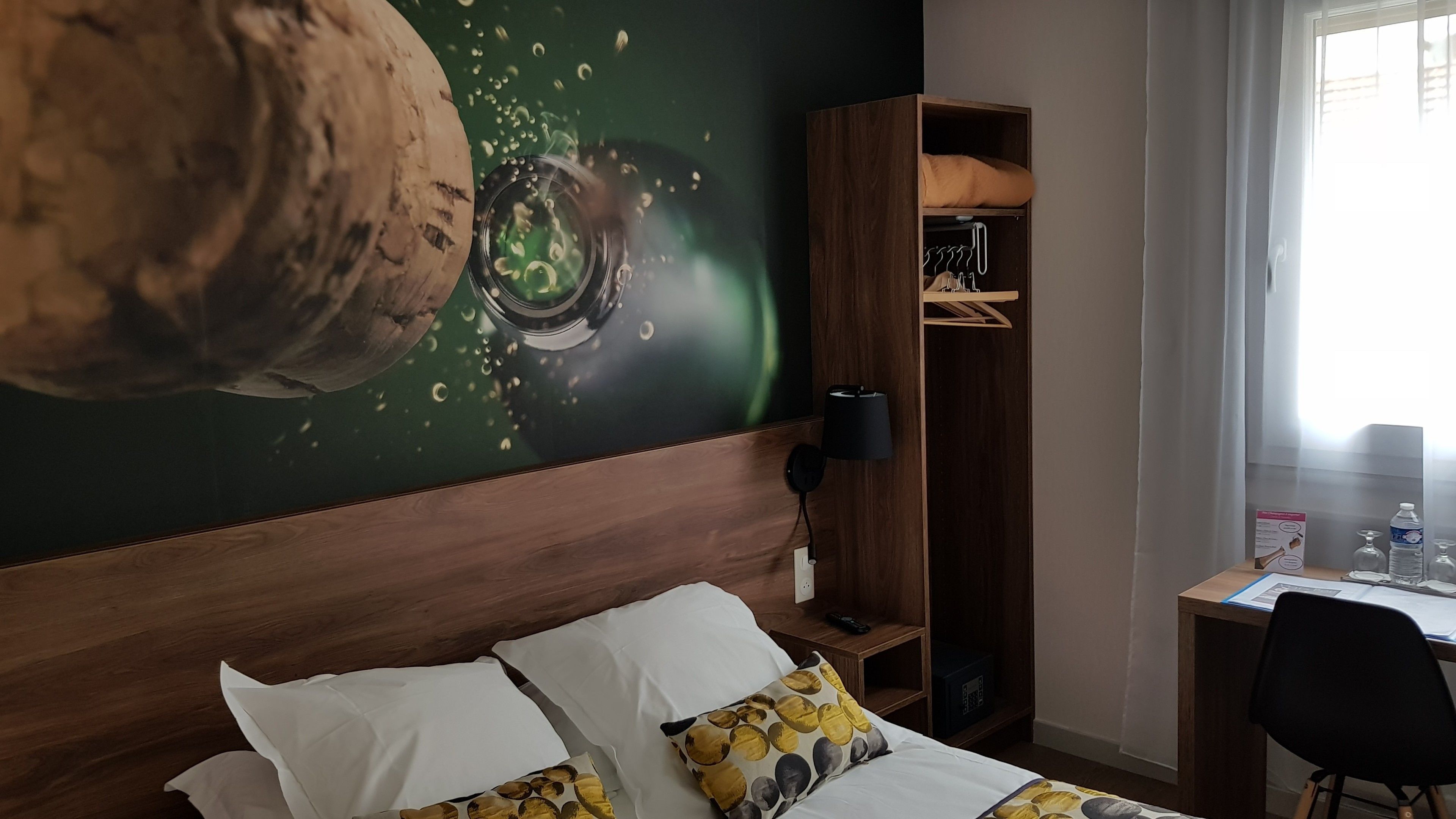 A hotel room with a large wall mural, a wooden wardrobe, a desk with a chair, and a bed with pillows.