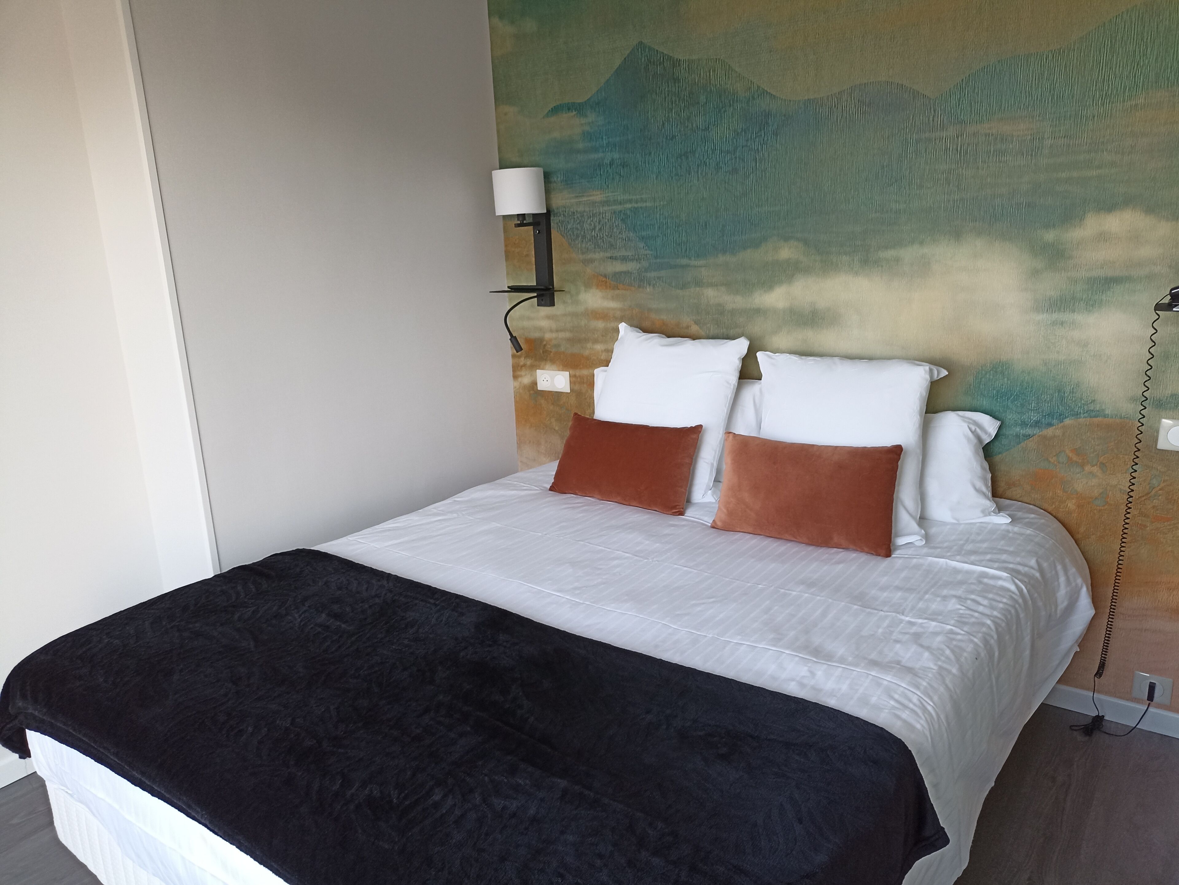 Una cama bien hecha con almohadas blancas y marrones, una manta de color negro y una lámpara de pared.
