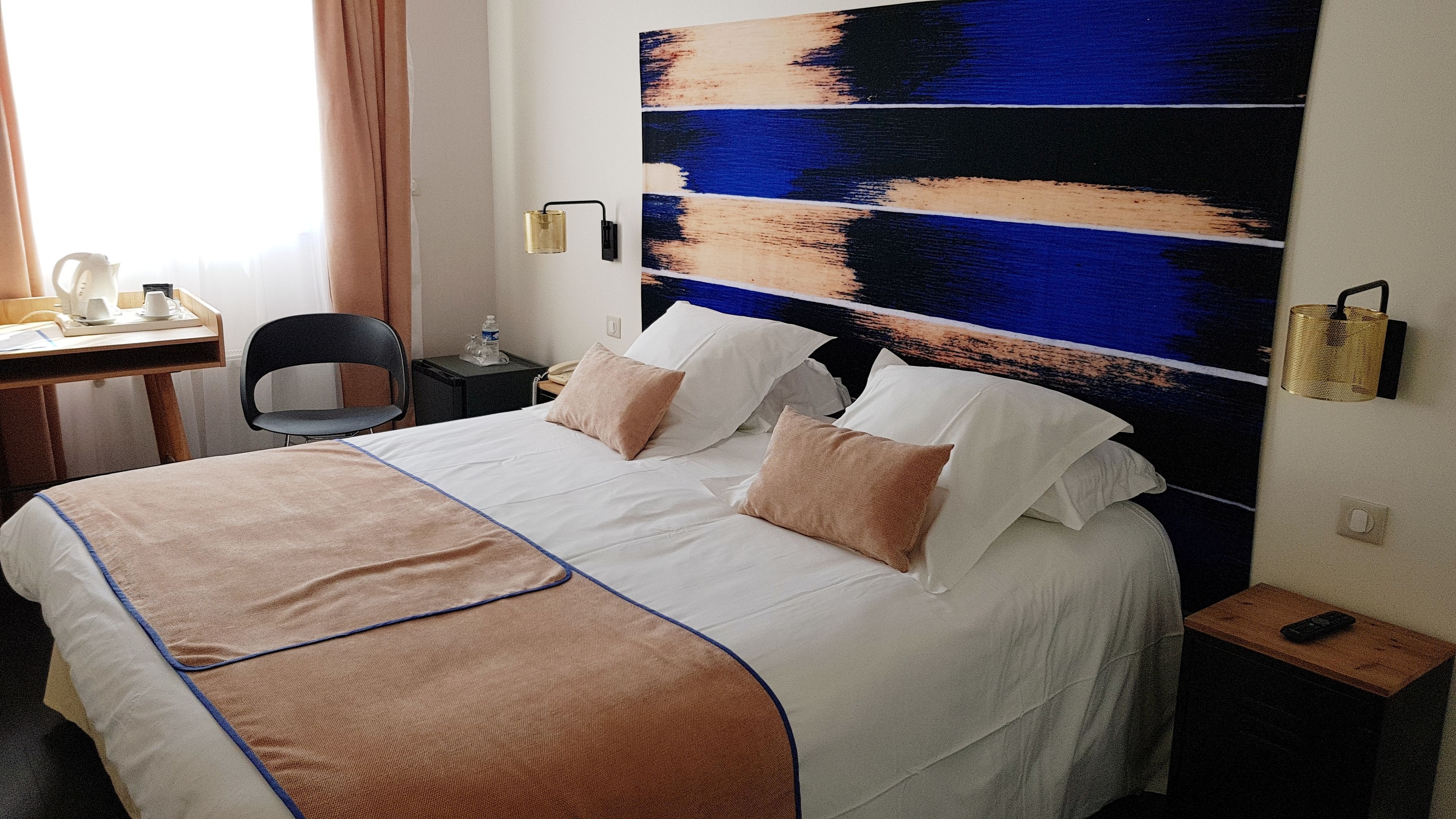 A hotel room with a bed, bedside tables, a desk with a chair, and a wall with a blue and beige abstract painting.