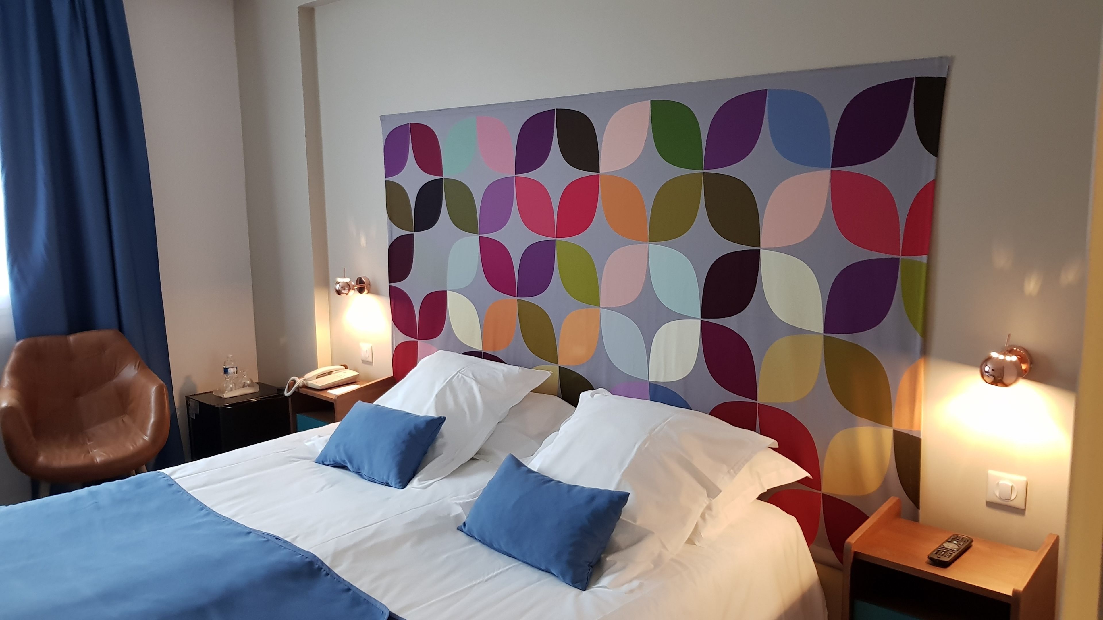 Een hotelkamer met een groot bed, twee kussens, een nachtkastje, een lamp, een stoel en een kleurrijk kunstwerk aan de muur.