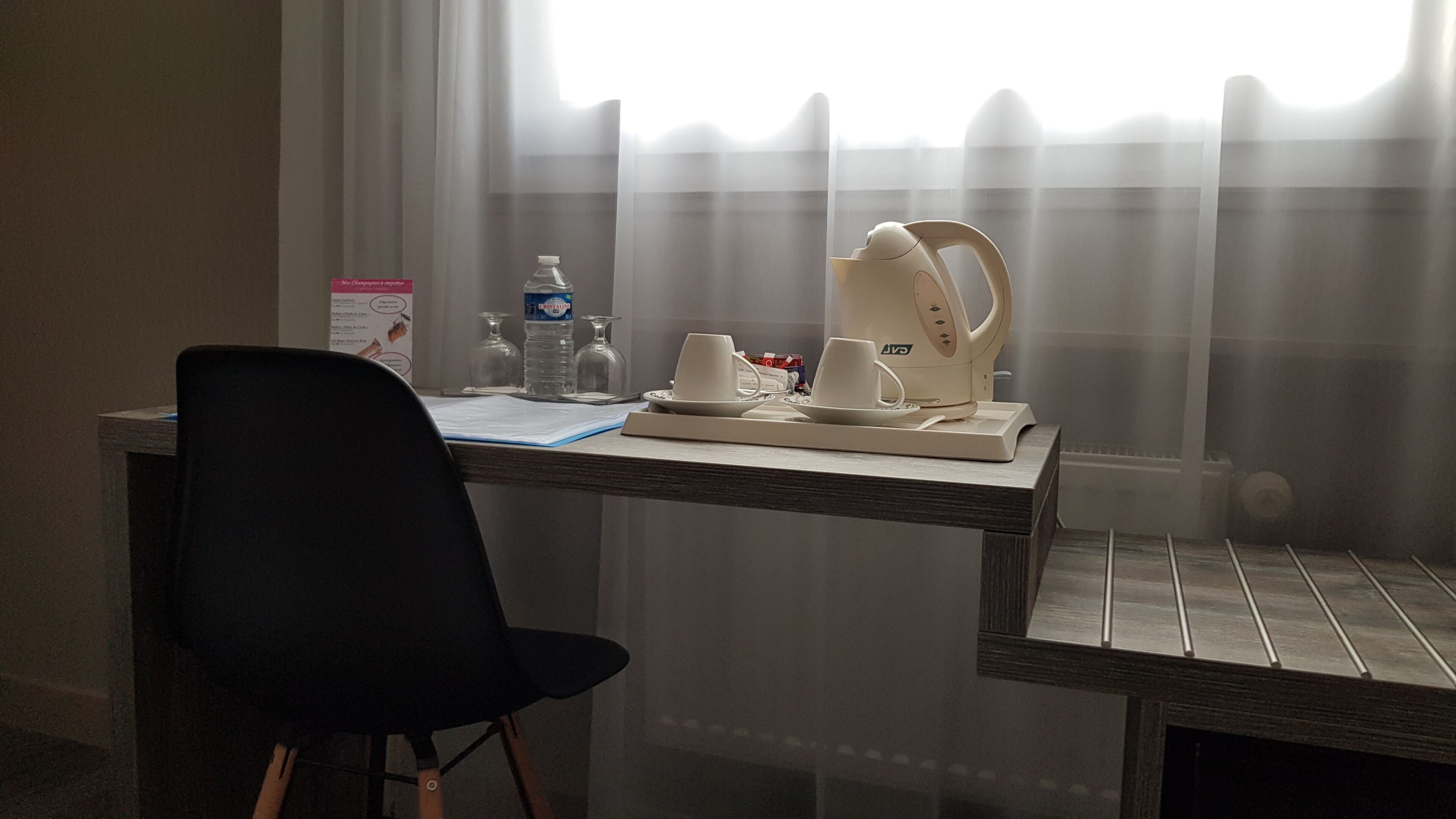 A hotel room with a wooden desk, a black chair, a coffee maker, cups, a water bottle, and a glass.