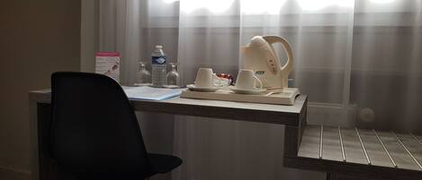 A hotel room with a wooden desk, a black chair, a coffee maker, cups, a water bottle, and a glass.